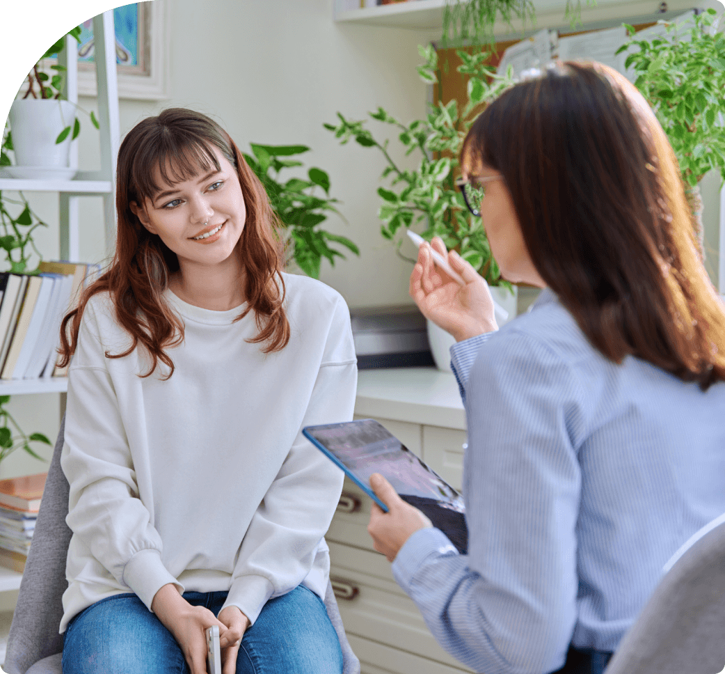 Two people sit in a serene room; one with a notebook, discussing health and wellness, while the other listens attentively. Shelves adorned with books and lush plants create a calming backdrop.