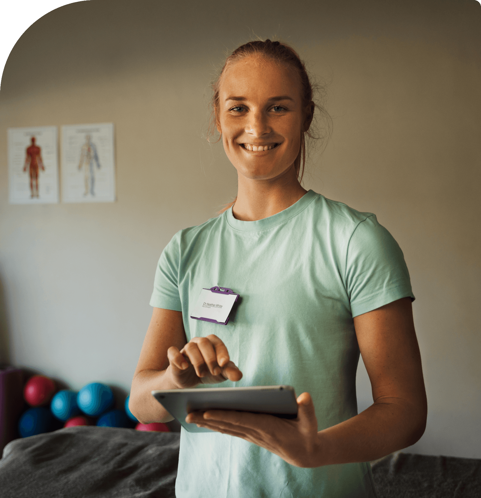 Person in a mint green shirt smiling and holding a tablet in a room adorned with anatomy posters and exercise balls, exuding an aura of health and wellness.